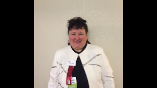 A woman in a white jacket and black shirt is standing in front of a white wall.