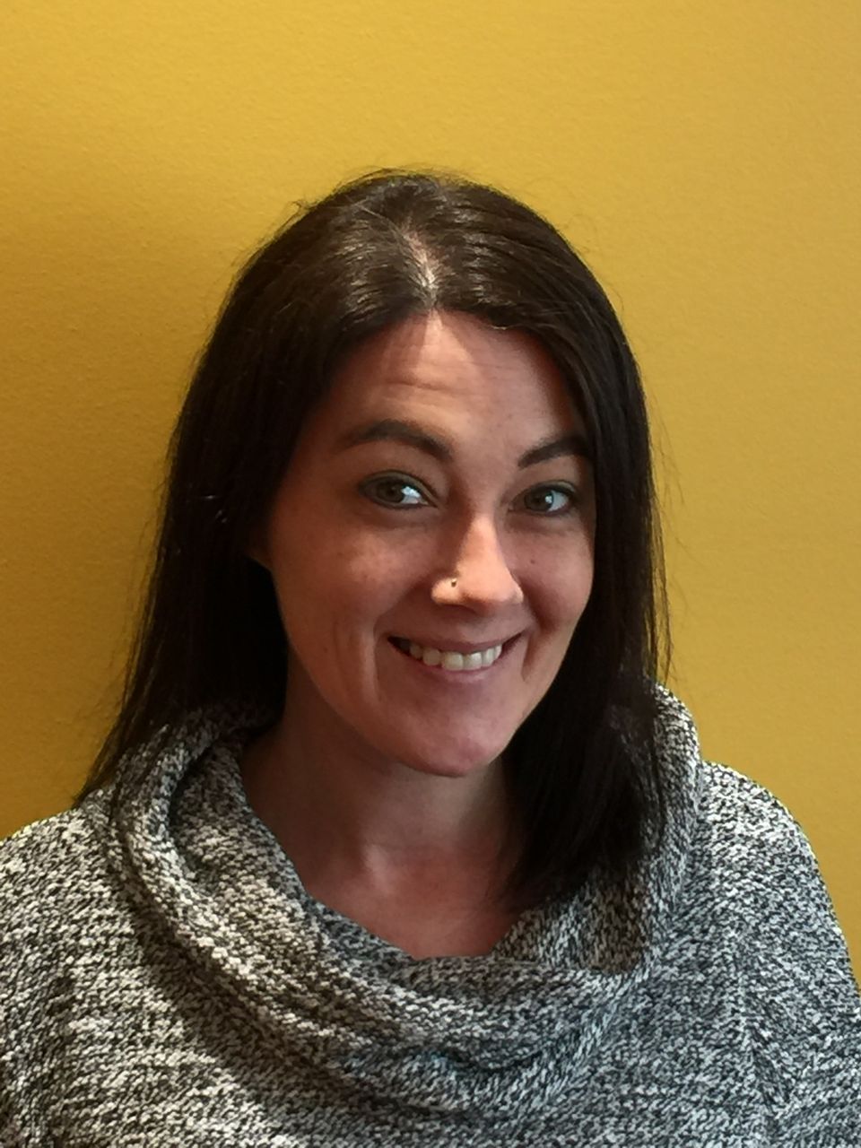 A woman wearing a grey sweater is smiling in front of a yellow wall.