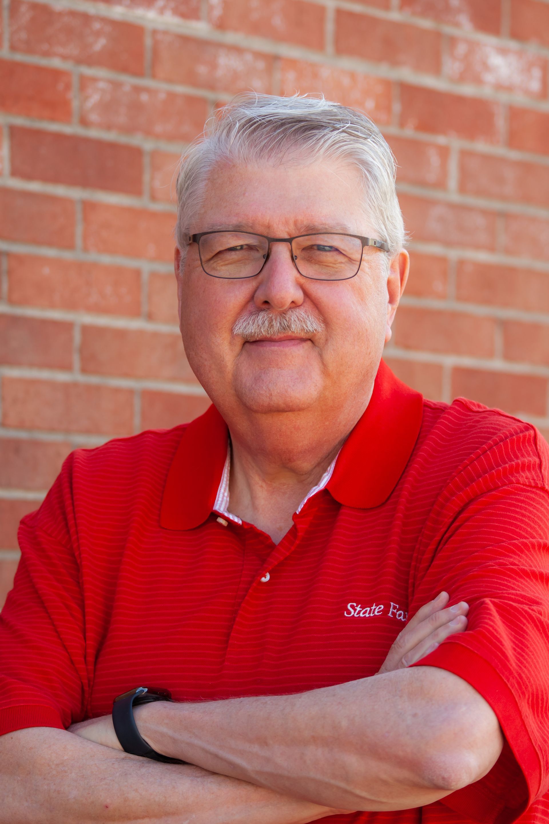 A man wearing glasses and a red shirt is standing in front of a brick wall with his arms crossed.