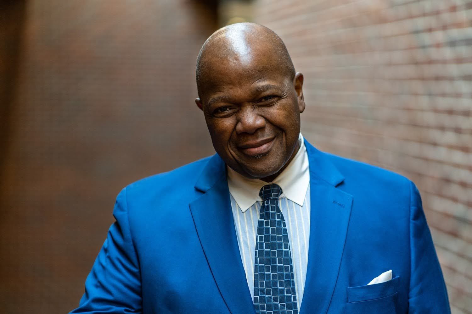 A man in a blue suit and tie is standing in front of a brick wall.