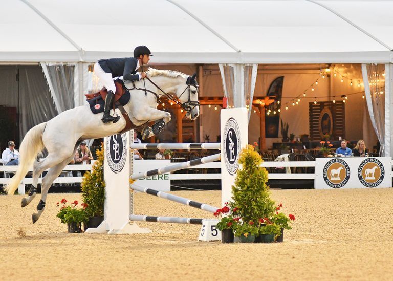 A man is riding a white horse over a jump.