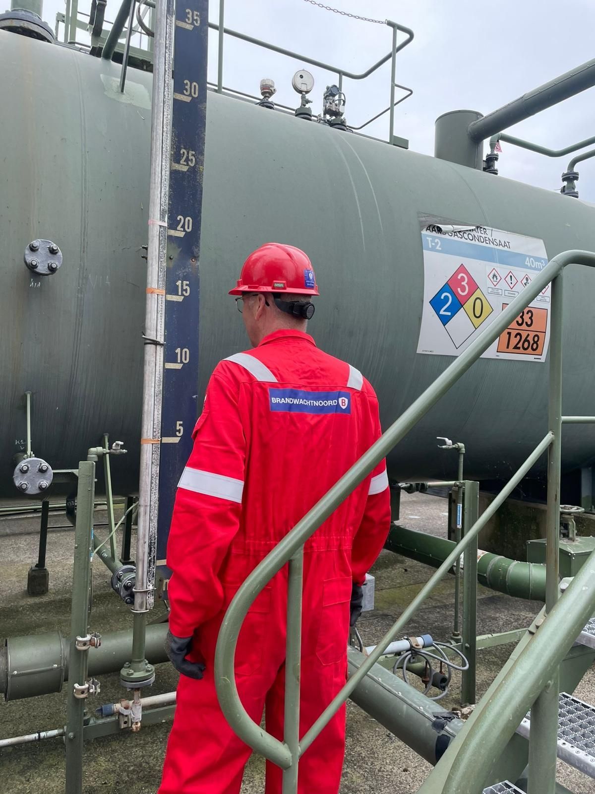 Een industriële brandwacht in een rode overall en veiligheidshelm staat naast een grote industriële tank met leidingen en veiligheidsborden. Op chemieparken of deGasunie en de NAM