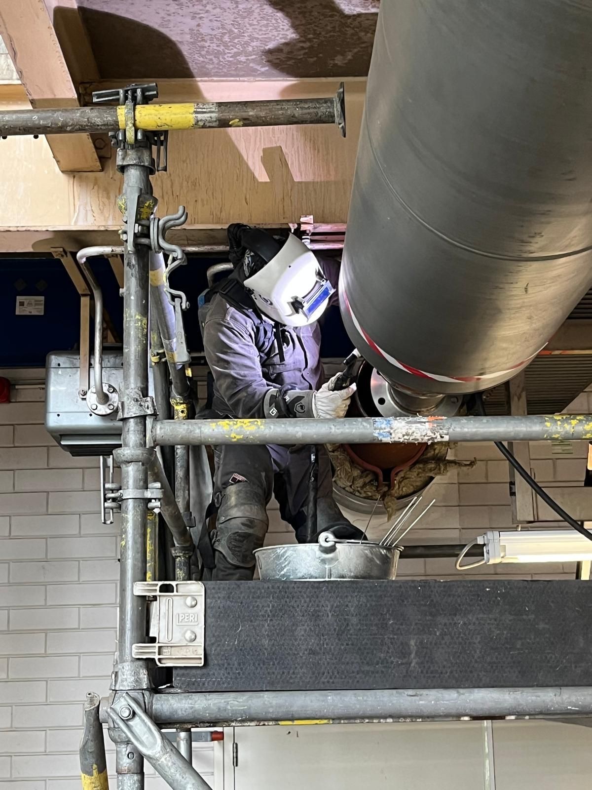 Een werknemer in beschermende kleding last pijpen in een industriële ruimte, met grote luchtkanalen boven zijn hoofd.