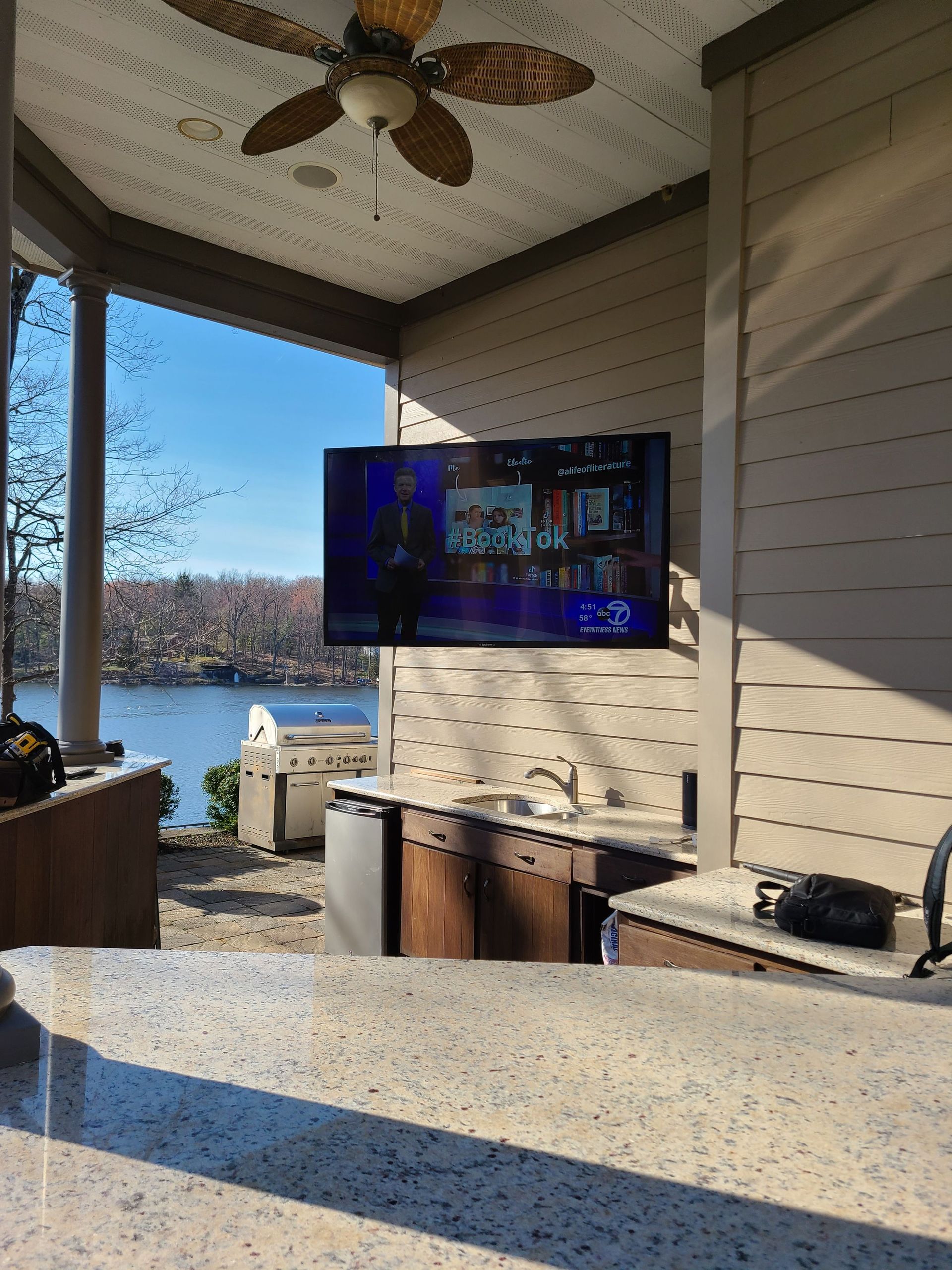 Covered patio overlooking water with wall-mounted TV, ceiling fan, and grill station by Audio Video Concepts