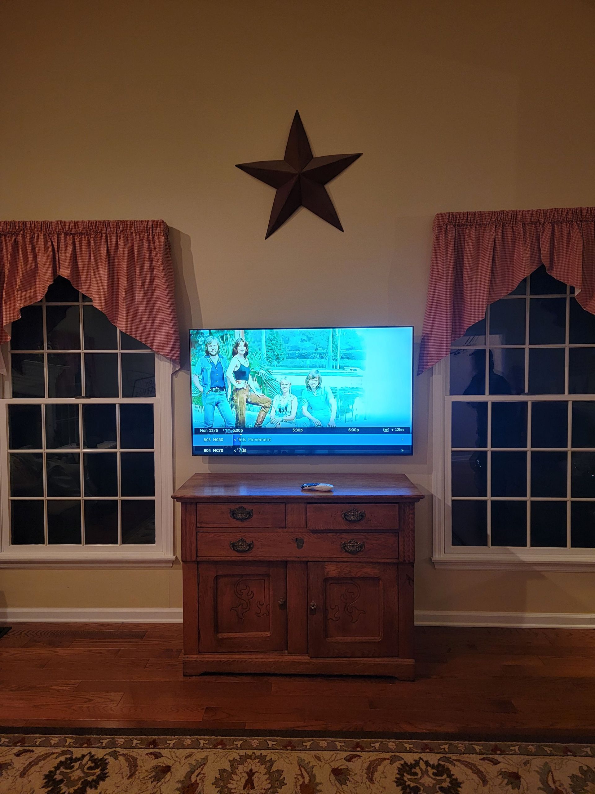 TV on a wooden cabinet between two windows with red curtains and a star wall decoration above by Audio Video Concepts