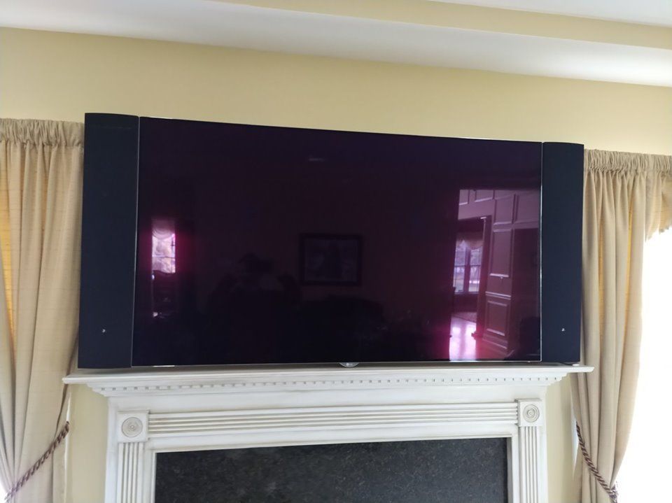 Large flat-screen TV above a white fireplace with beige curtains by Audio Video Concepts