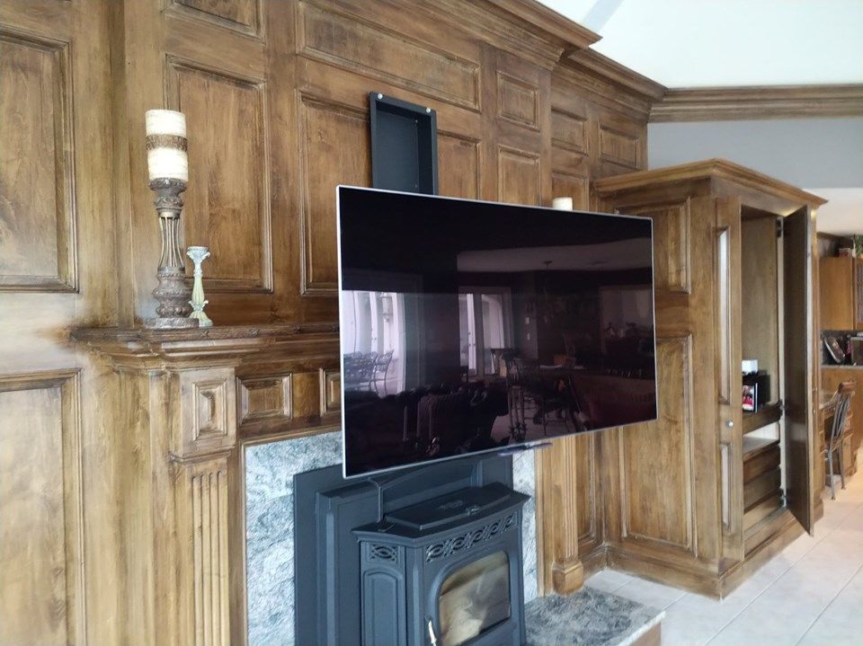 Large flat-screen TV mounted above a black wood stove in a wood-paneled room by Audio Video Concepts