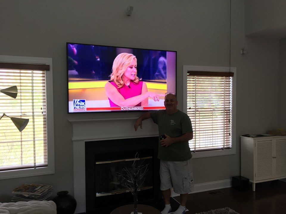 Person standing in a living room beside a wall-mounted TV showing a blonde woman on a news program Audio Video by Concepts