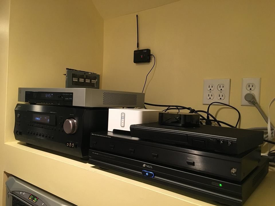 Black audio equipment stacked on a shelf with tangled cables and wall outlets behind it by Audio Video Concepts