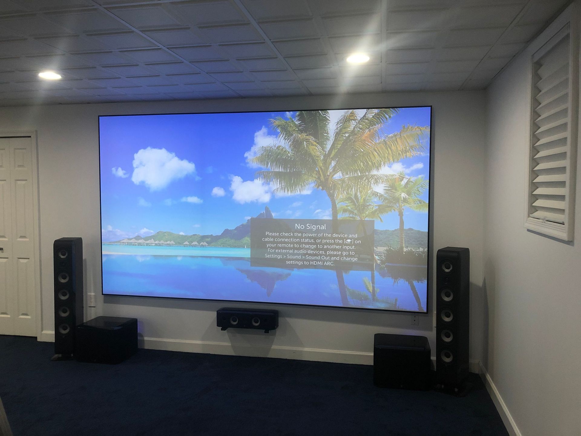Home theater room with large TV showing tropical beach scene, flanked by tall speakers by Audio Video Concepts