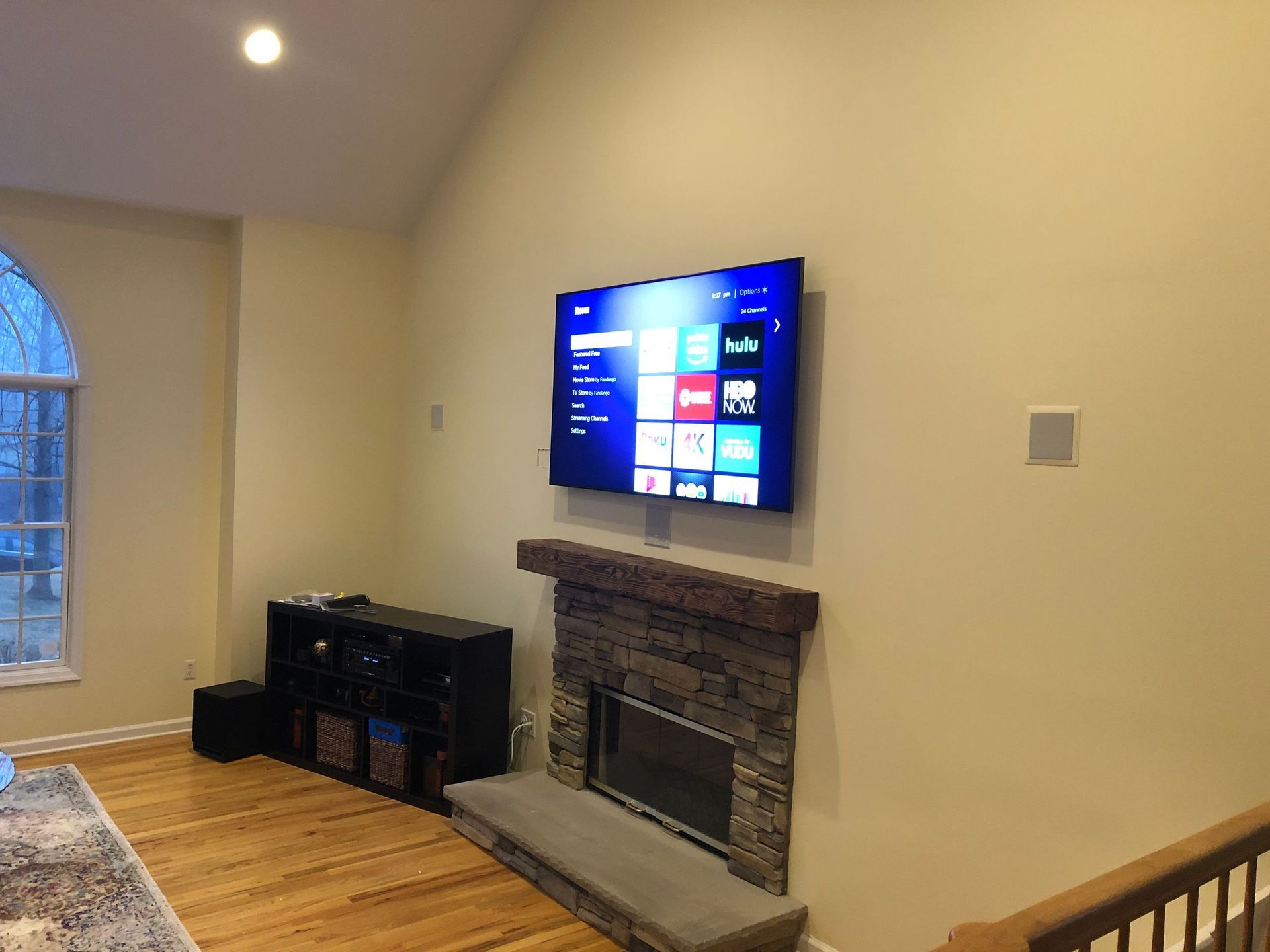 Living room with mounted TV above a stone fireplace, hardwood floor, and arched window by Audio Video Concepts