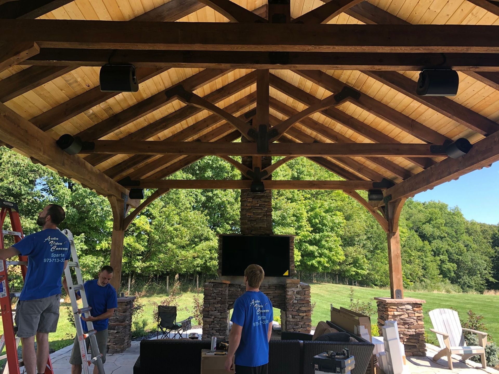 Open-air pavilion with exposed wood beams, stone fireplace, and people standing inside outdoors by Audio Video Concepts