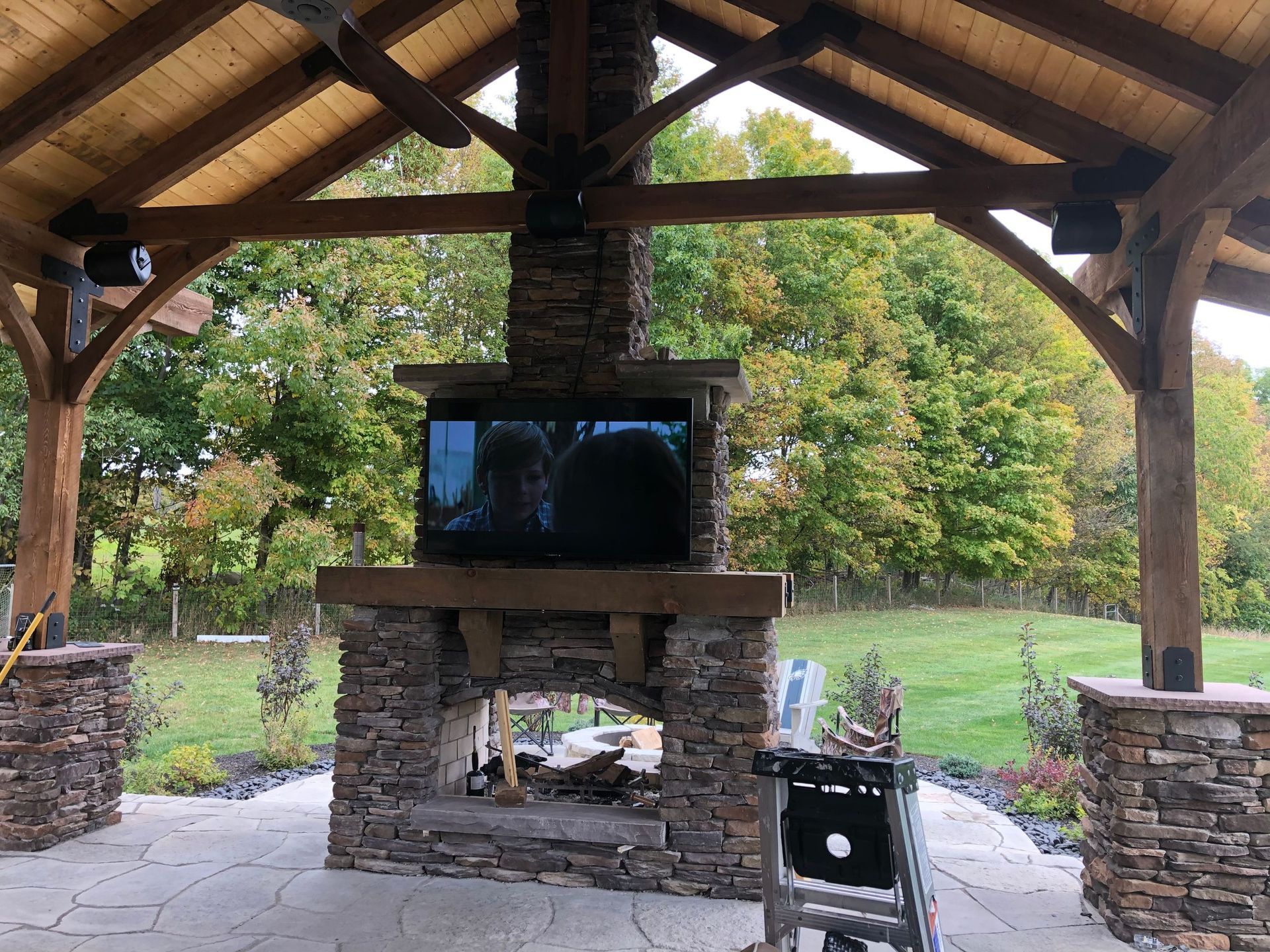 Covered outdoor stone fireplace under a wooden pavilion in a grassy yard by Audio Video Concepts