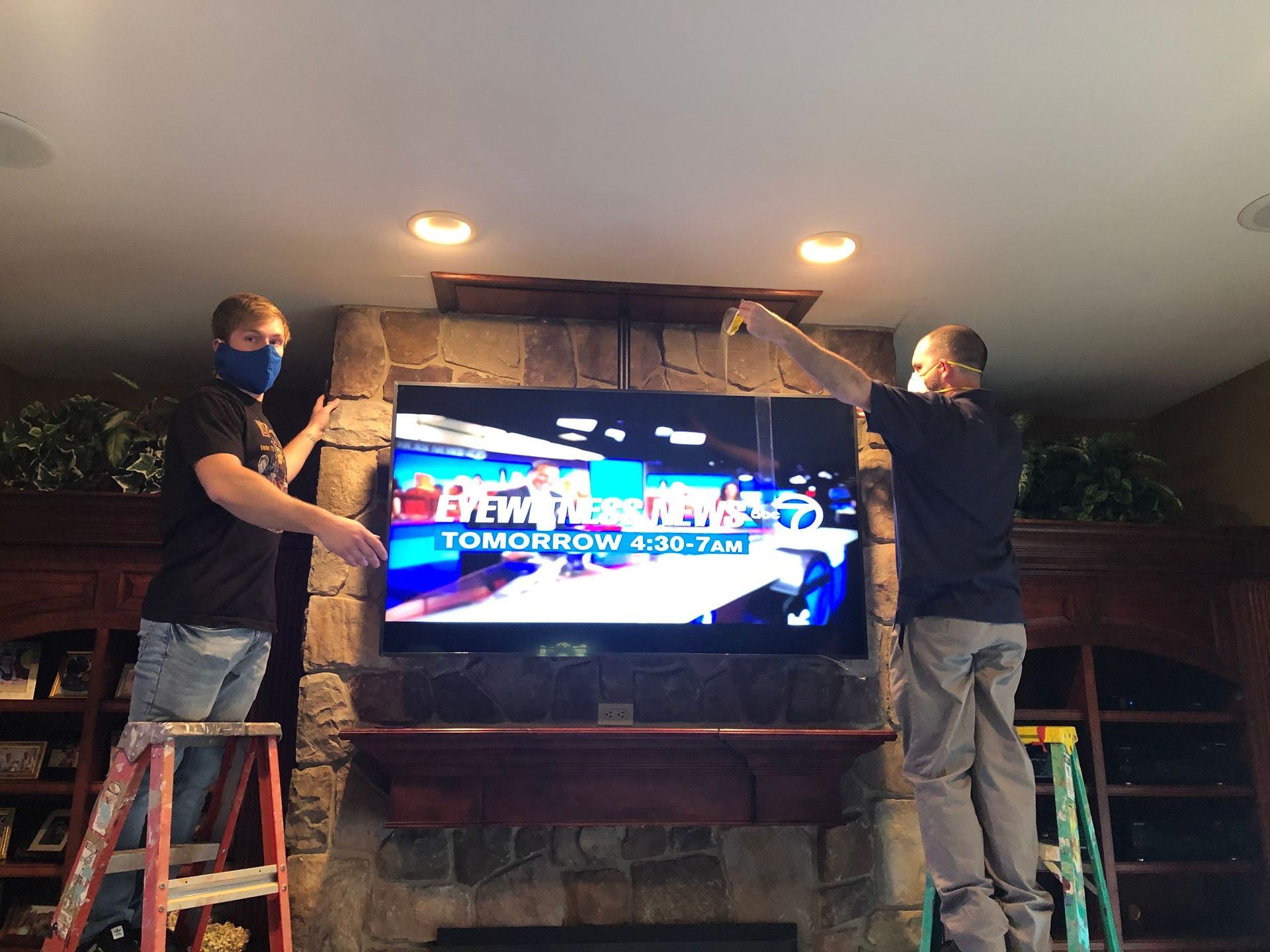 Two people on ladders mount a large TV into a stone fireplace mantel by Audio Video Concepts
