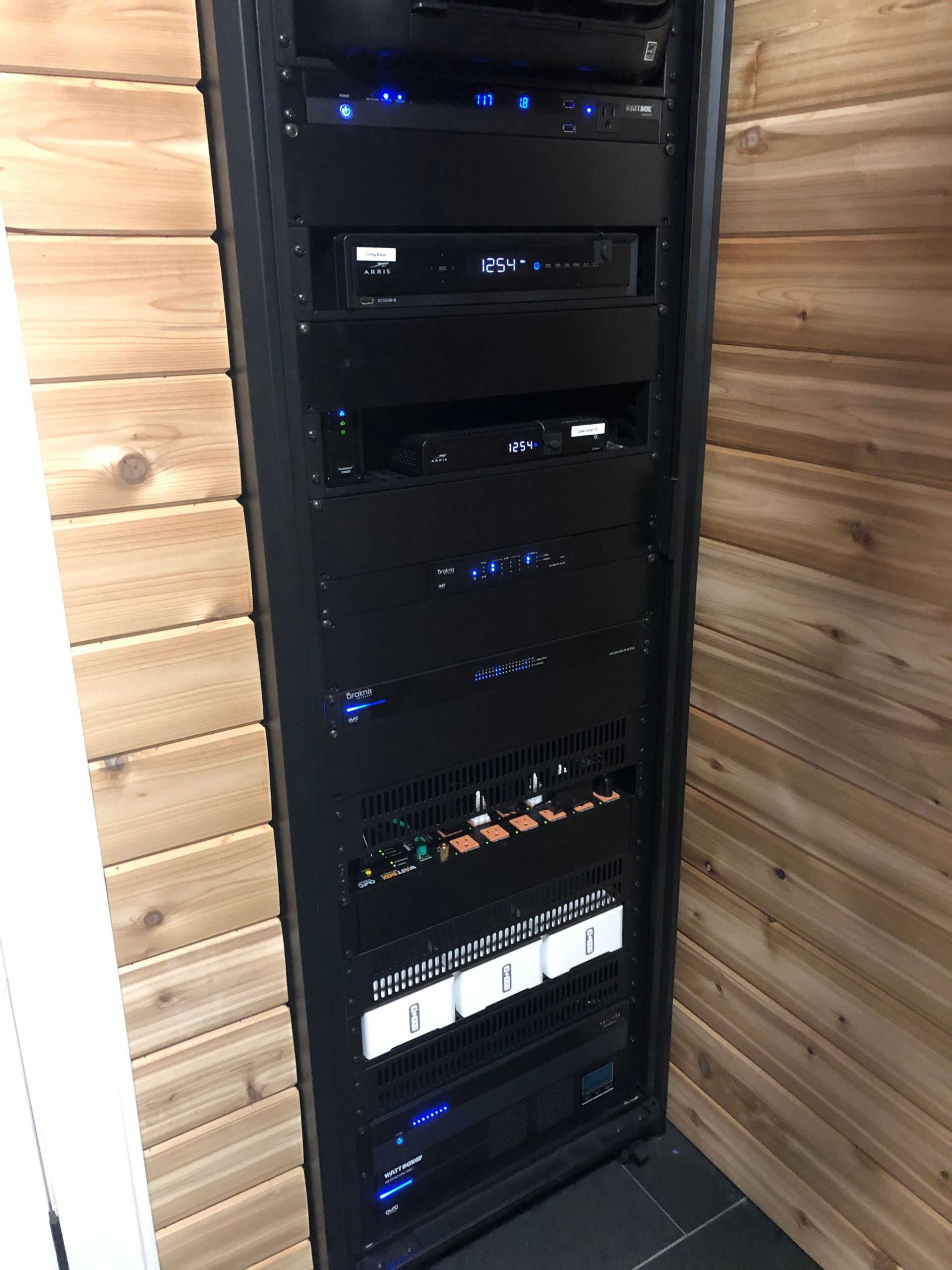 Tall black equipment rack with audio/video components in a wood-paneled corner closet by Audio Video Concepts