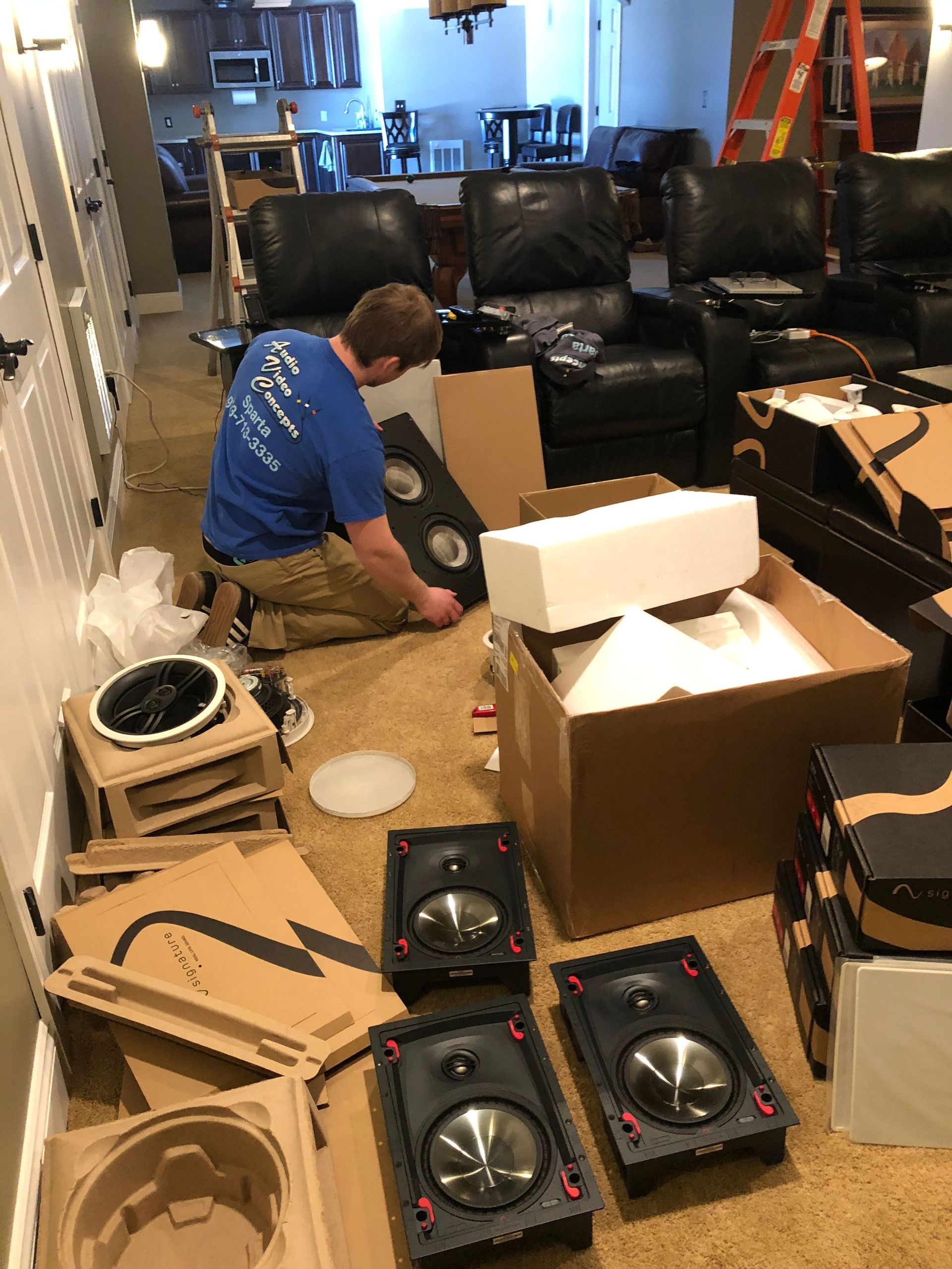 Person kneeling on a living room floor amid boxes, tools, and speakers during home setup or unpacking by Audio Video Concepts