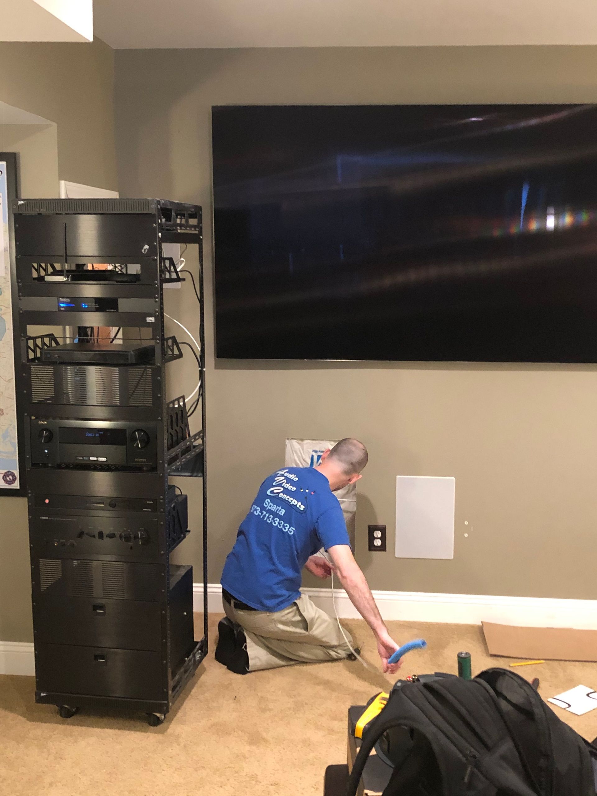 Technician kneeling by a wall under a large TV, working with cables beside audio equipment racks by Audio Video Concepts