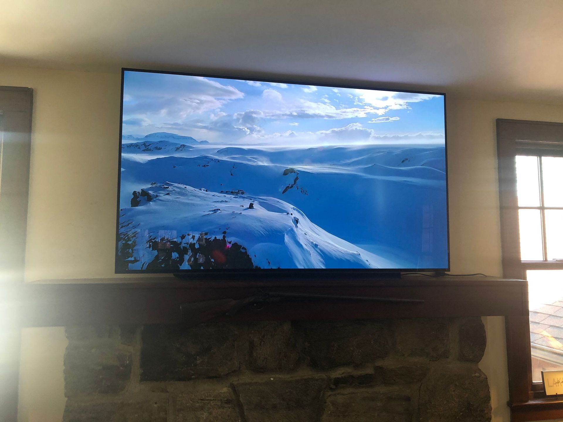 Large TV above a stone fireplace displaying a snowy mountain ski view in a bright living room by Audio Video Concepts