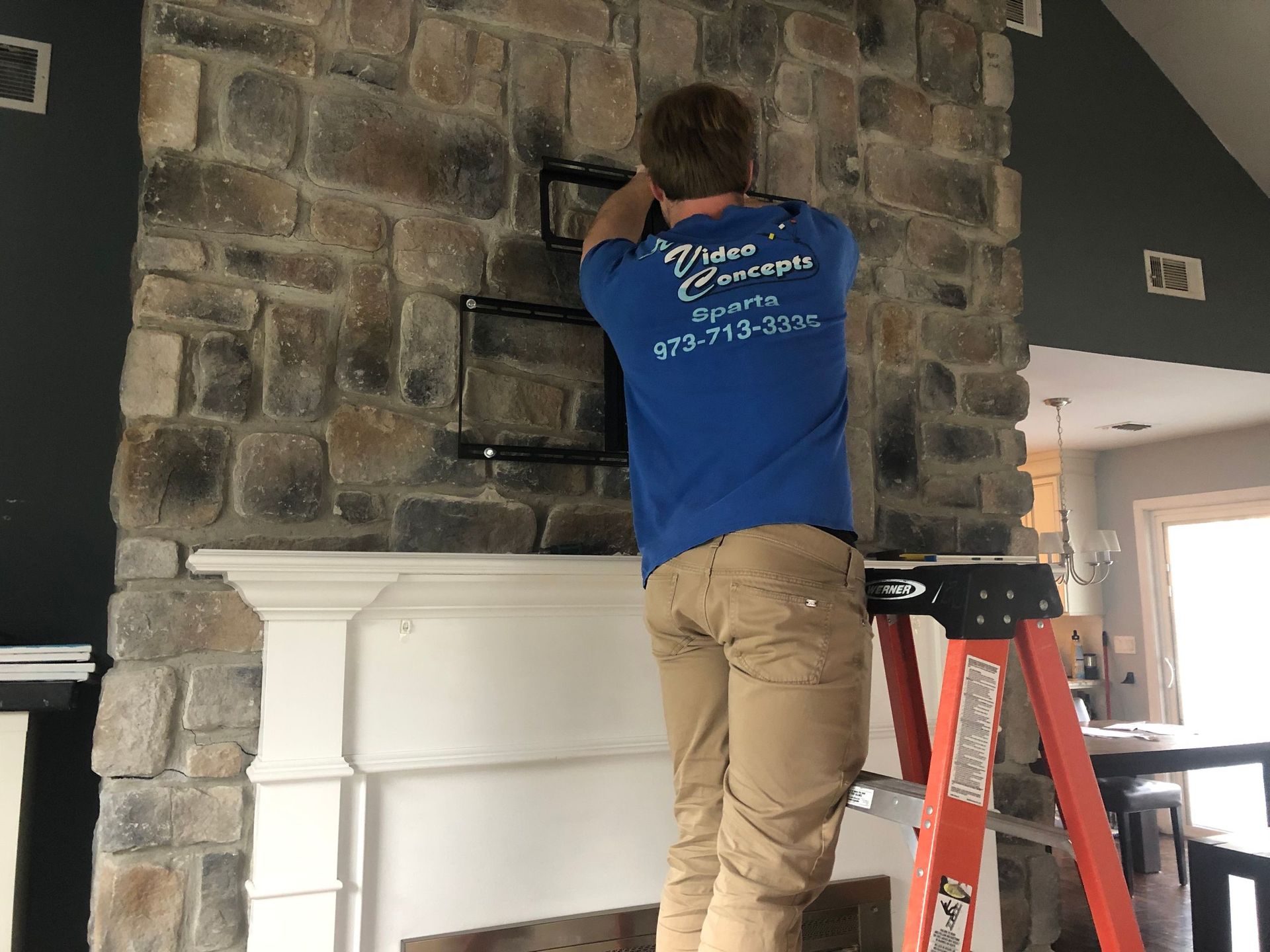 Person in a blue shirt works on a stone fireplace beside a red ladder in a home interior by Audio Video Concepts