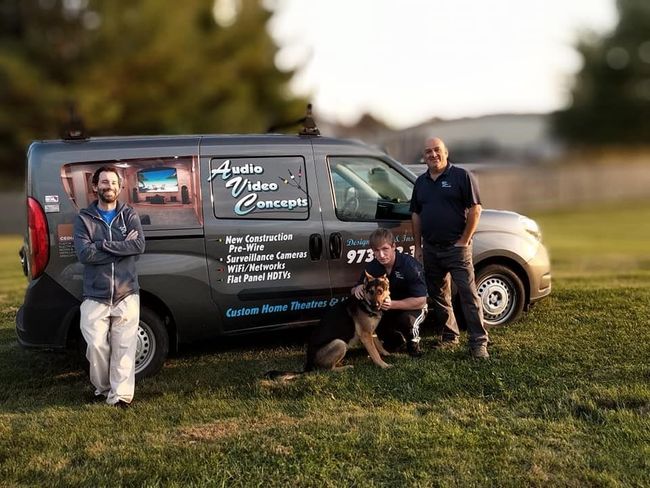 Three people posing with a gray van on a grassy field, one holding a dog by Audio Video Concept