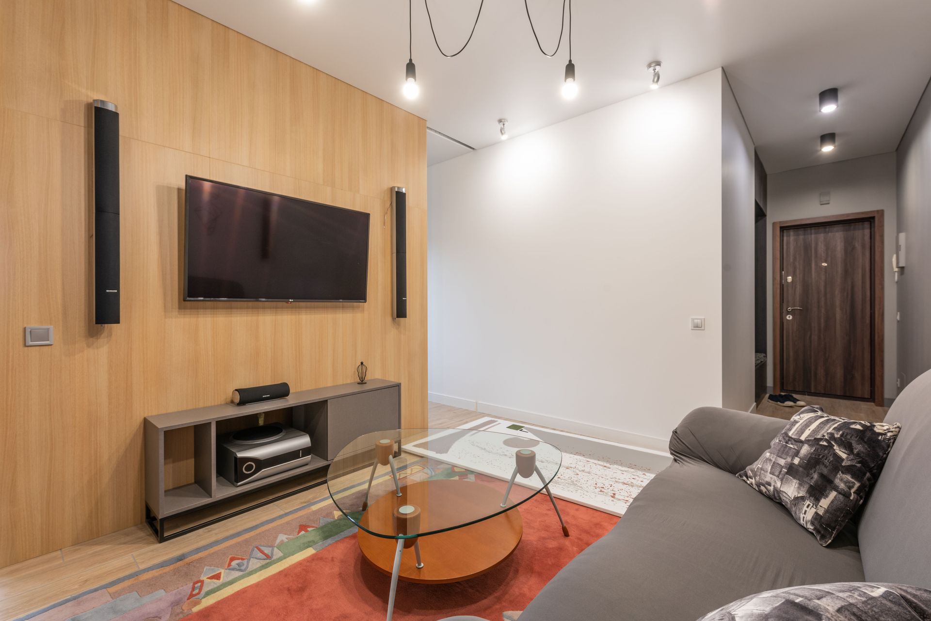 Modern living room with wall-mounted TV, gray sofa, glass coffee table, and wood accent wall by Audio Video Concepts