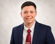 Studio headshot of a smiling person in a navy suit, white shirt, and red tie on a light background