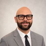 Portrait of a bald man with glasses and a beard in a gray suit and striped tie