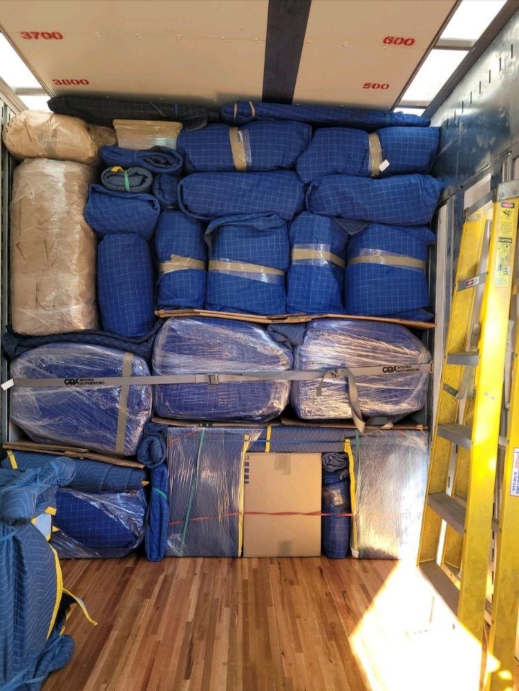 Cargo van interior loaded with stacked blue and beige wrapped packages, strapped down with yellow tie-downs.