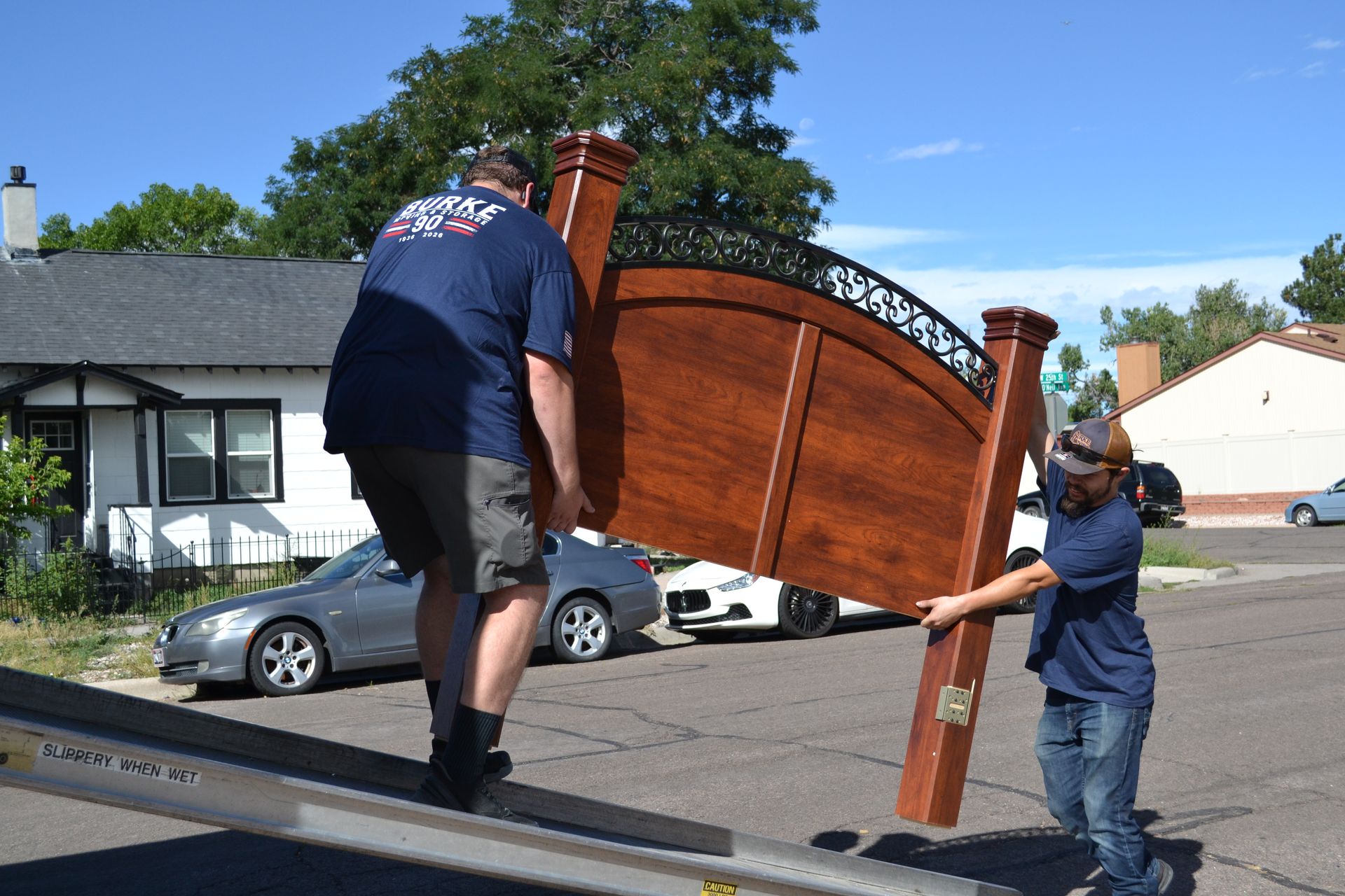 Two people lifting a large wooden bed frame onto a truck ramp outdoors