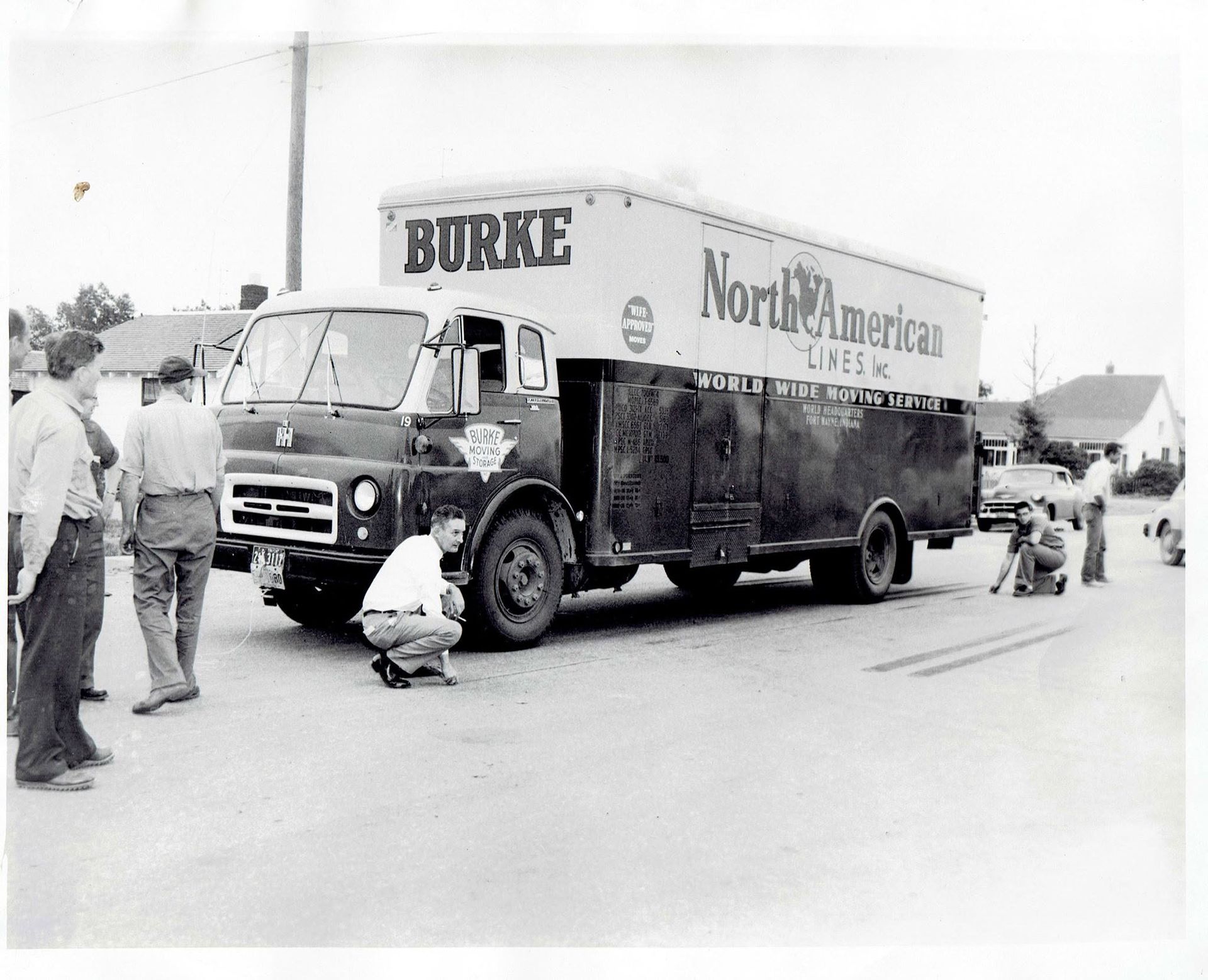 Two men kneel beside a North American truck parked on a street during a roadside service stop.