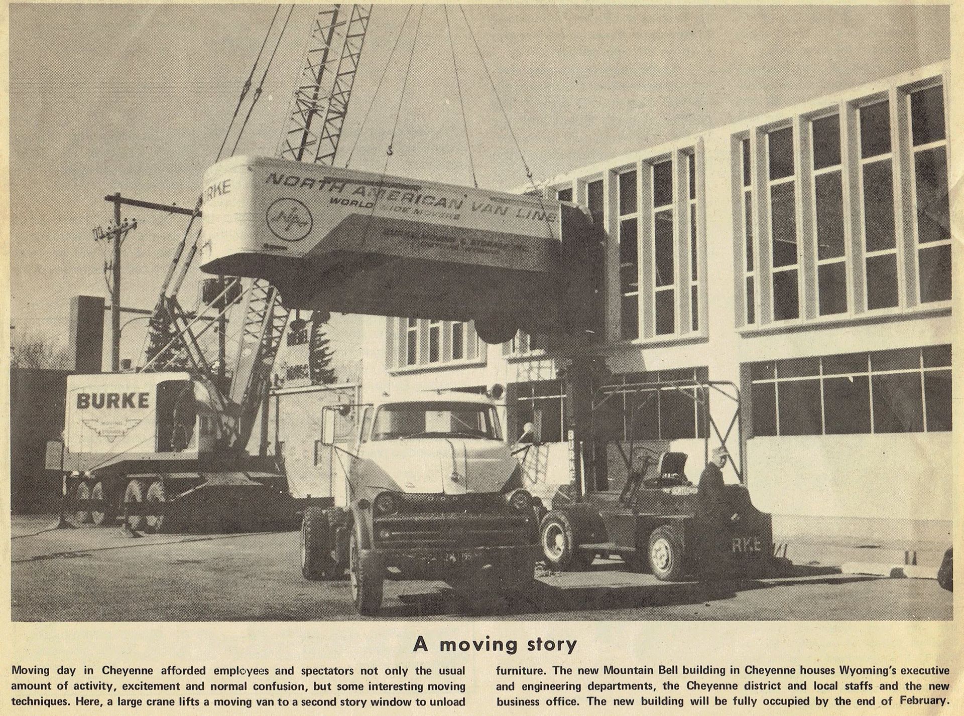 Crane lifting a large concrete beam over trucks beside a building.