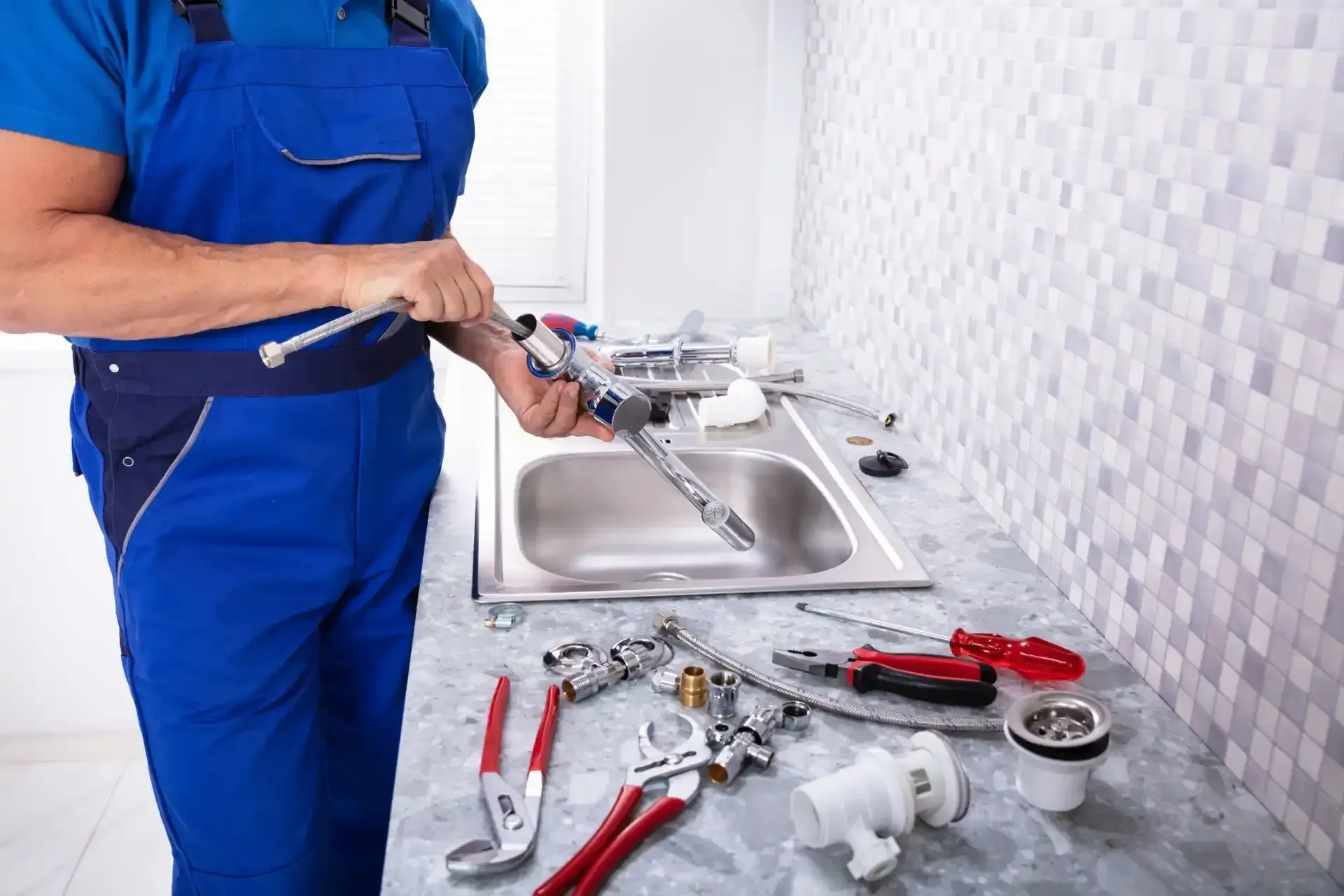 Plumber in blue overalls repairing a sink faucet with tools spread on the counter