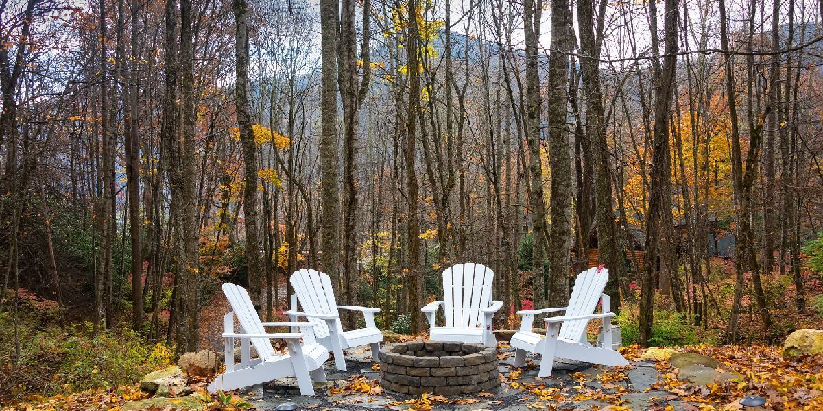 Rustic forest landscape with white Adirondack chairs arranged around a stone fire pit in autumn.