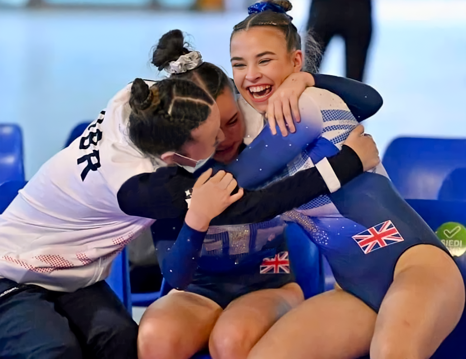 Gymnasts in blue and white leotards embrace, smiling, with a coach in a 