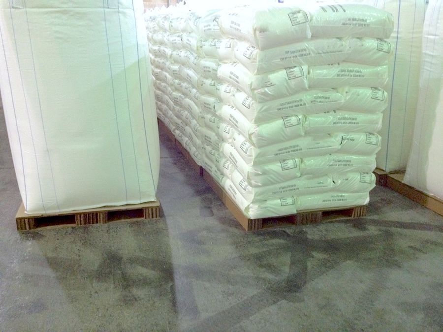 Large white bulk bag and stacked bags of chemical material on corrugated pallets inside a manufacturing facility.