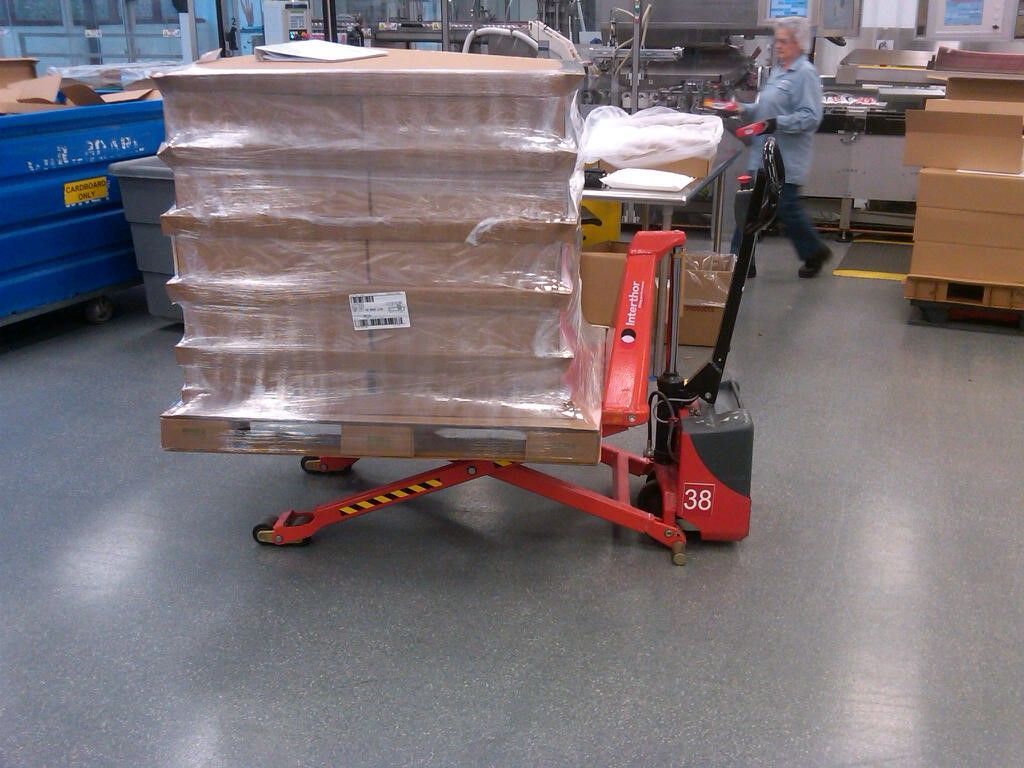 Shrink-wrapped cartons on a corrugated pallet deck raised by a scissor-lift pallet jack inside a manufacturing facility.
