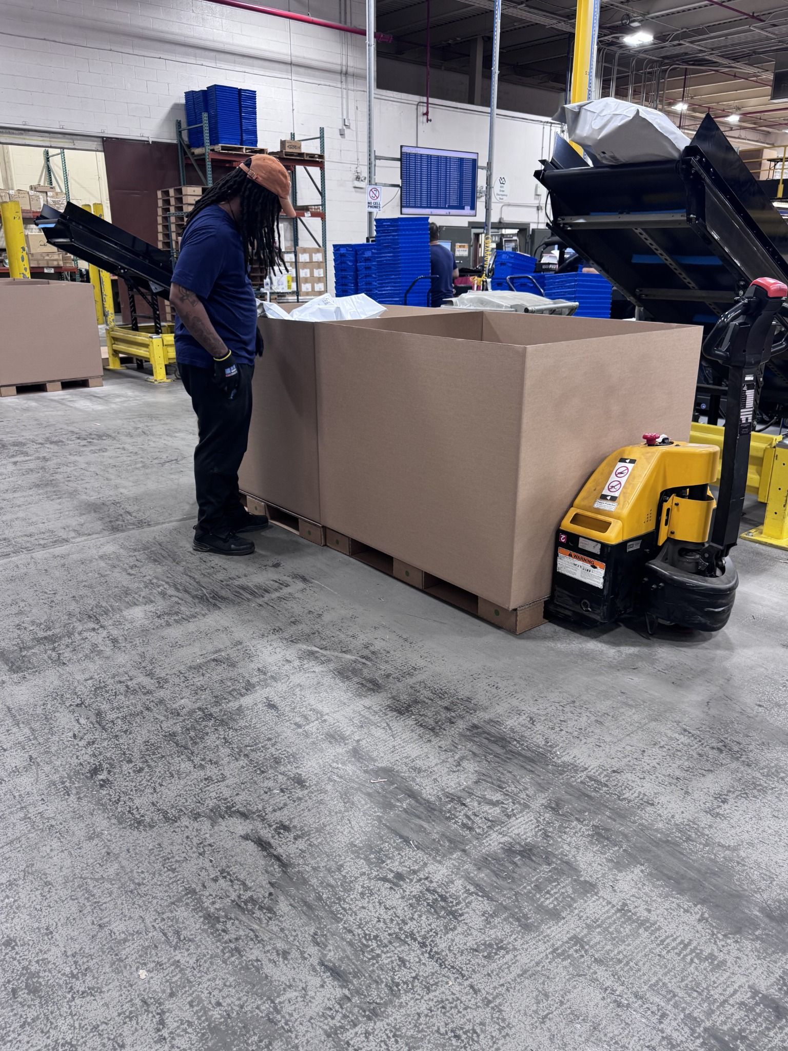 Warehouse employee working beside an open bulk container on a corrugated pallet, with powered material-handling equipment.