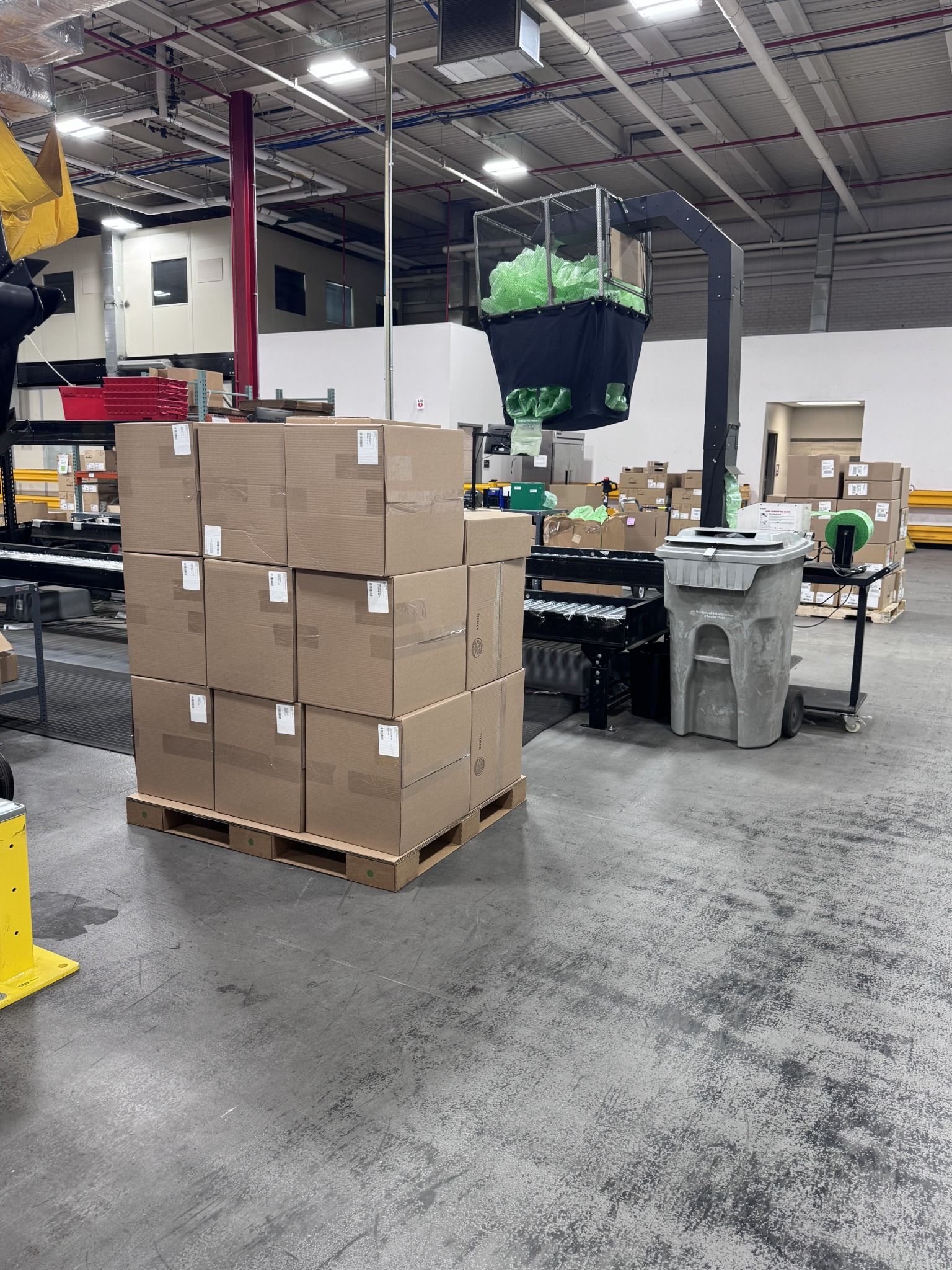 Cartons stacked on an Ecodek corrugated pallet beside automated packaging and conveyor equipment in a distribution facility.