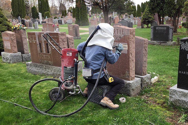 Person cleaning a gravestone in a cemetery with a sandblasting machine.