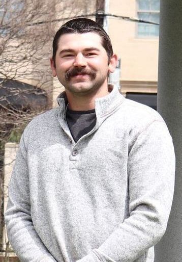 Man with mustache wearing a gray sweater, smiling in front of a building.