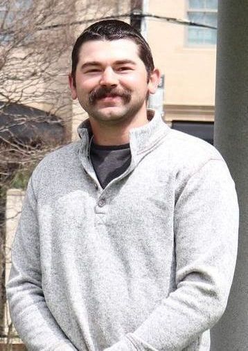 Man with mustache wearing a gray sweater, smiling slightly, outdoors.