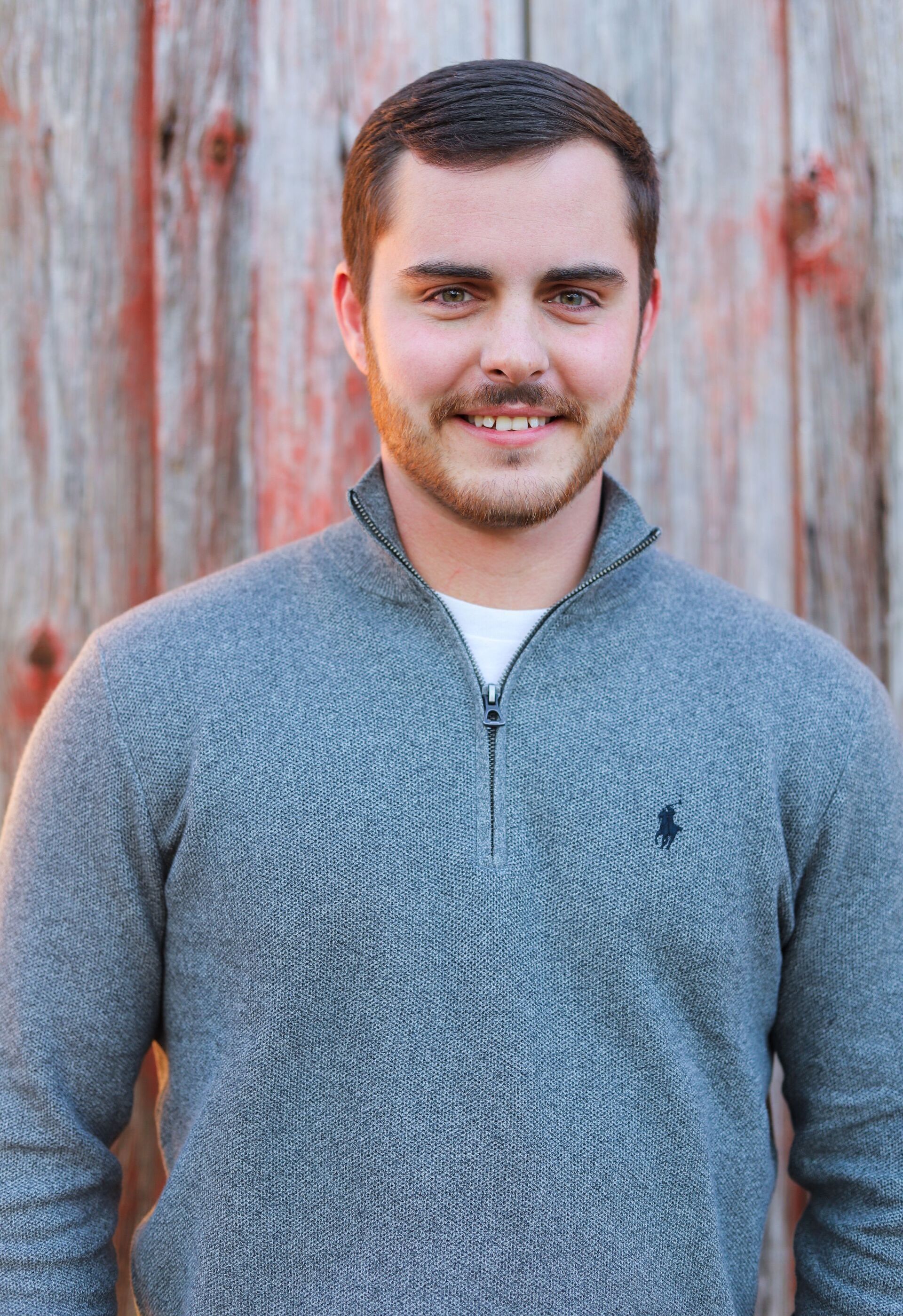 Man with short dark hair and stubble smiles, wearing gray sweater; red barn backdrop.