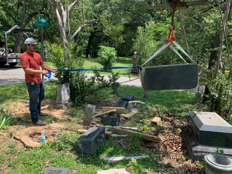 Man using a crane to lift a large dark tombstone in a cemetery on a sunny day.