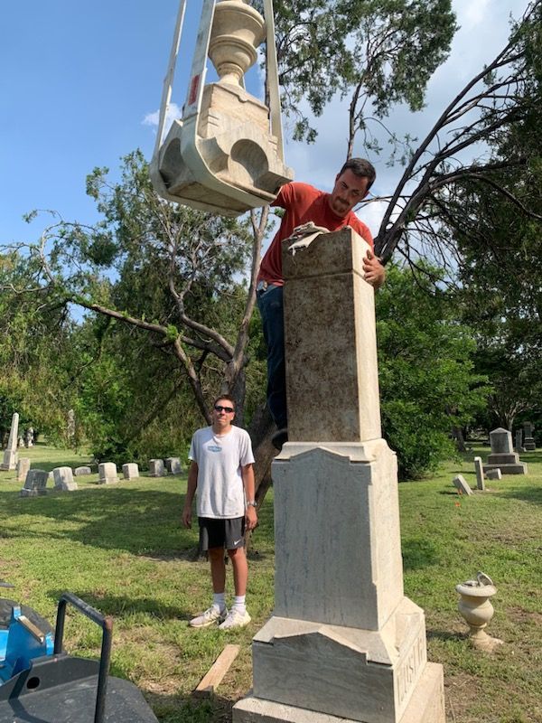Two men placing a large stone vase on top of a tombstone in a cemetery.