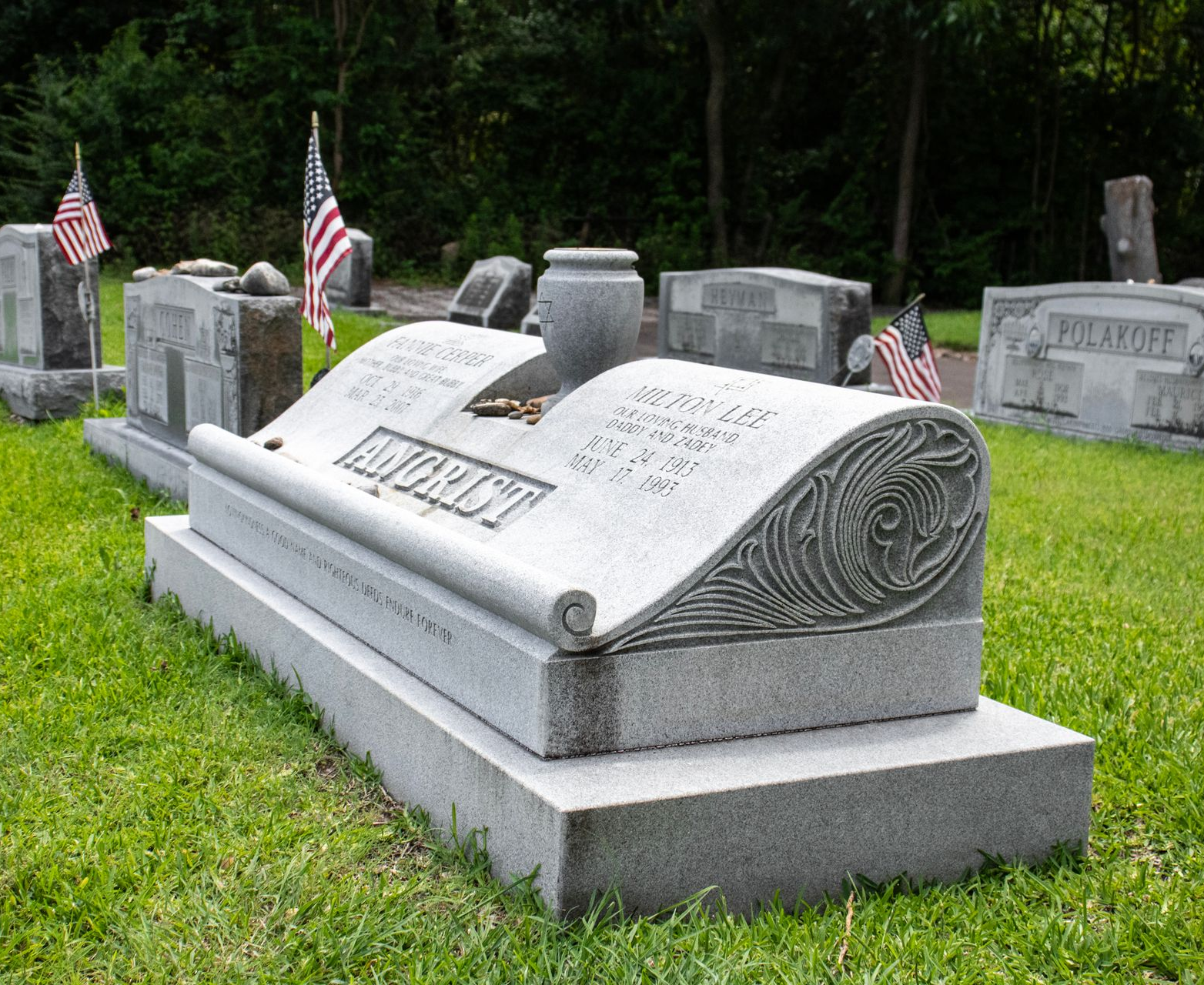 Gravestone with a scroll design in a grassy cemetery, flanked by American flags.