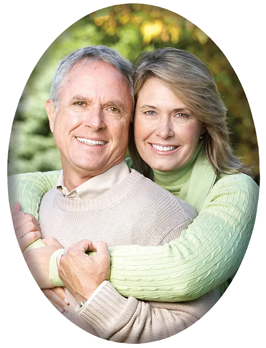 Couple smiling, embracing outdoors; woman's arms around man, green and beige clothing, natural background.