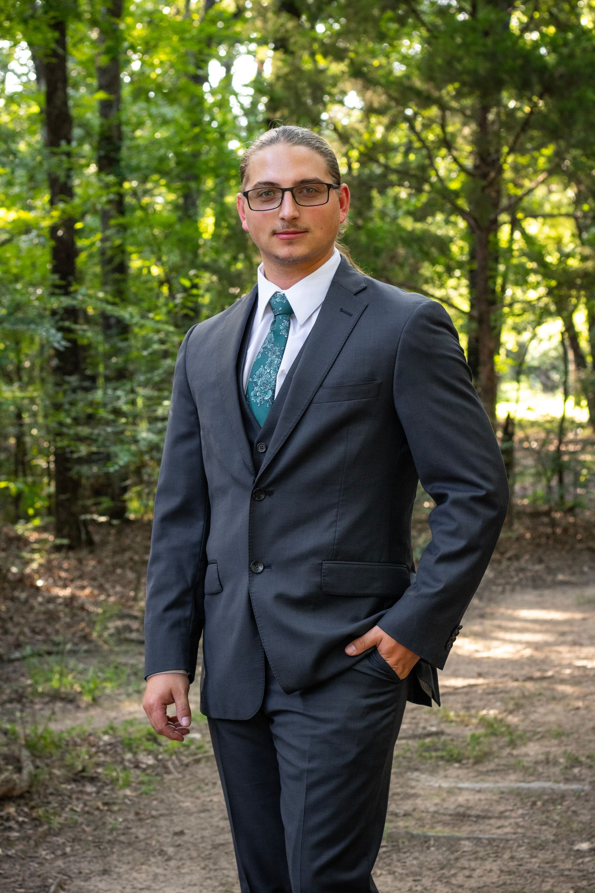 Man in a gray suit stands in a forest, hand in his pocket, looking forward.