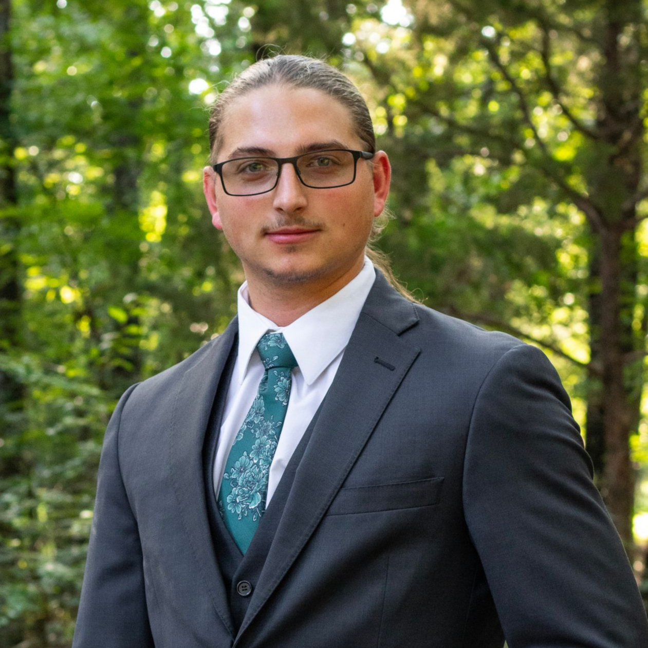 Man in suit and tie outdoors with glasses, trees in the background.