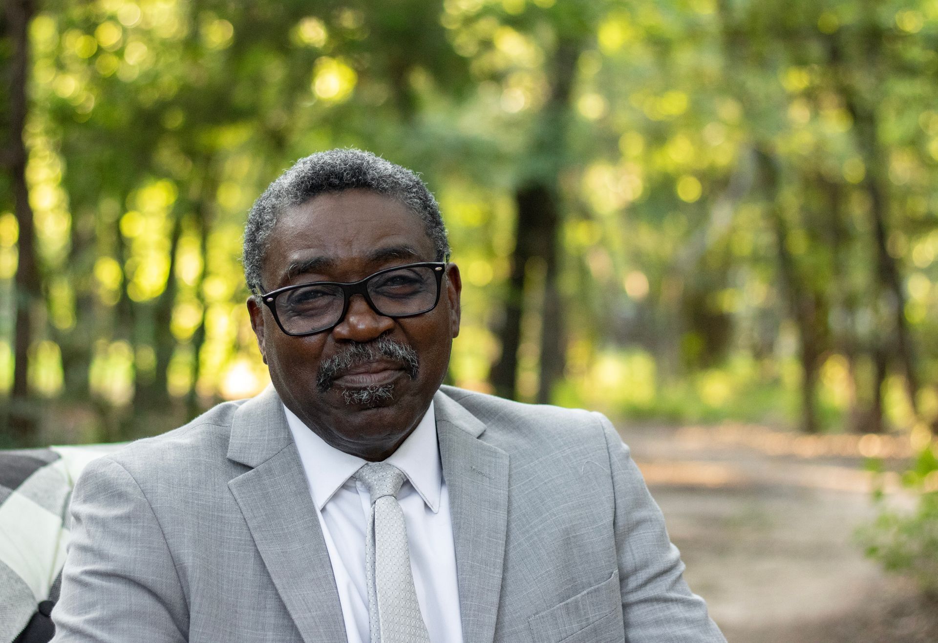 Man in glasses and gray suit, smiling outdoors with a green, blurred background.