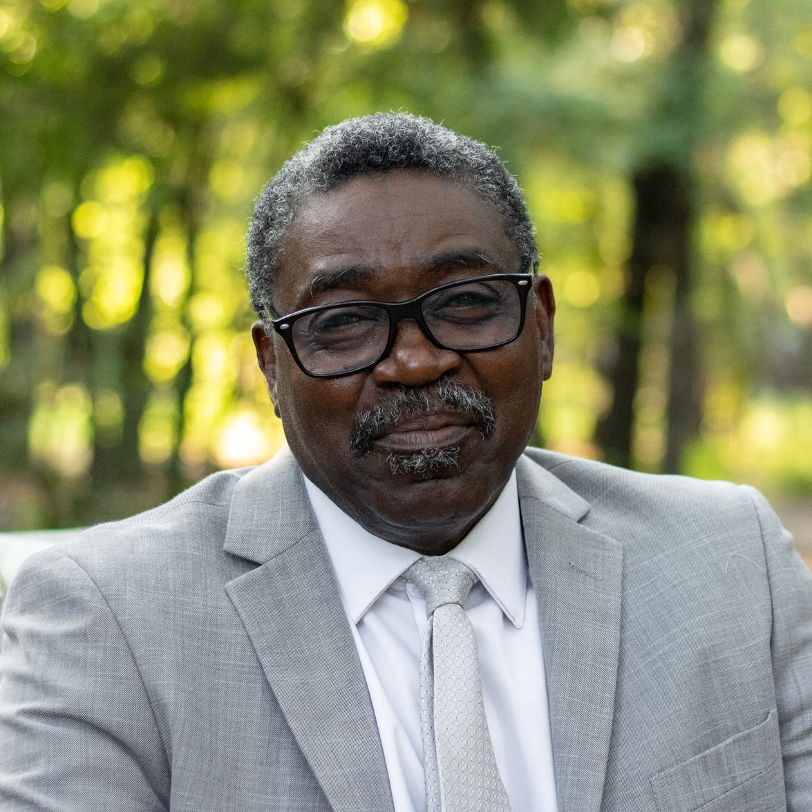 Man in a light gray suit and glasses, smiling. Outdoor setting with trees.